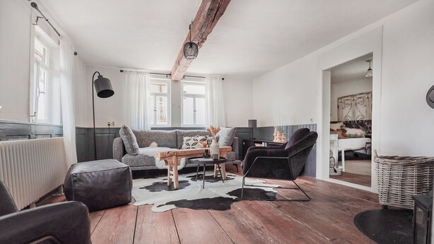 Uma sala de estar moderna com piso de madeira, um sofá cinza, uma poltrona preta e uma área de jantar com mesa e cadeiras.