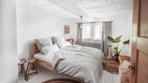 Quarto com cama de madeira, um banquinho pequeno, uma planta em vaso e janela com cortinas.