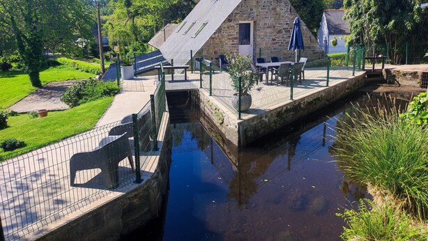 Een stenen gebouw met een schuin dak, een klein terras met tafels en stoelen, en een smalle waterloop ervoor.