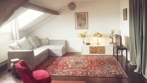 A cozy loft bedroom with a sloped ceiling, a white sofa, a red chair, a patterned rug, a bedside table with a lamp, and a framed picture on the wall. - Appartement 100m2 Dernier Étage Calme et Lumineux (Melun)