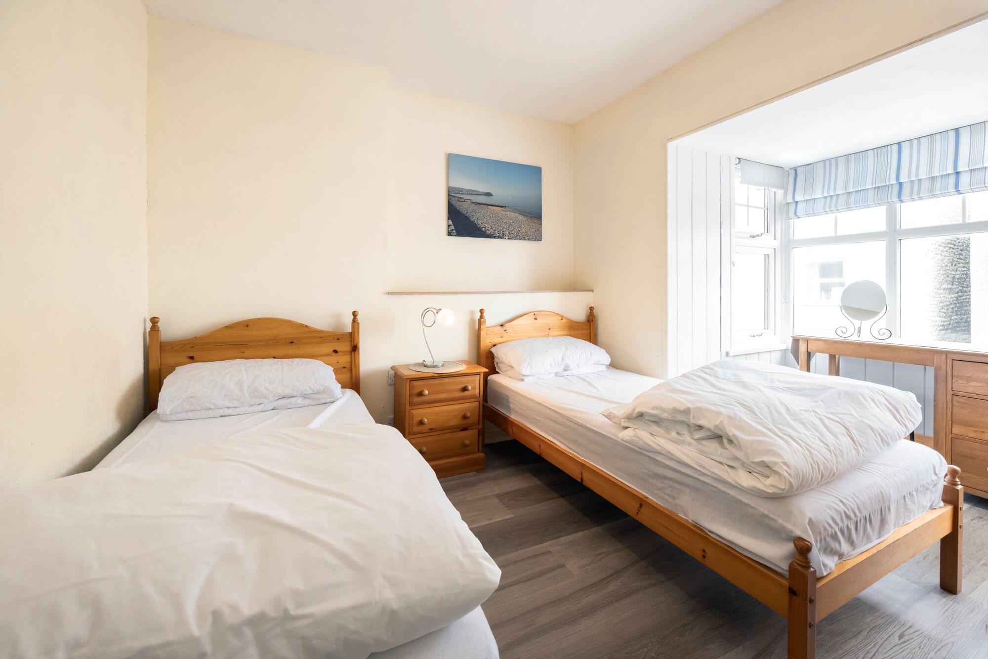 A bedroom with two single beds, a wooden dresser, a bedside table with a lamp, and a window with a view of the sea.