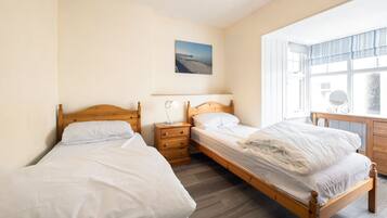 A bedroom with two single beds, a wooden dresser, a bedside table with a lamp, and a window with a view of the sea.