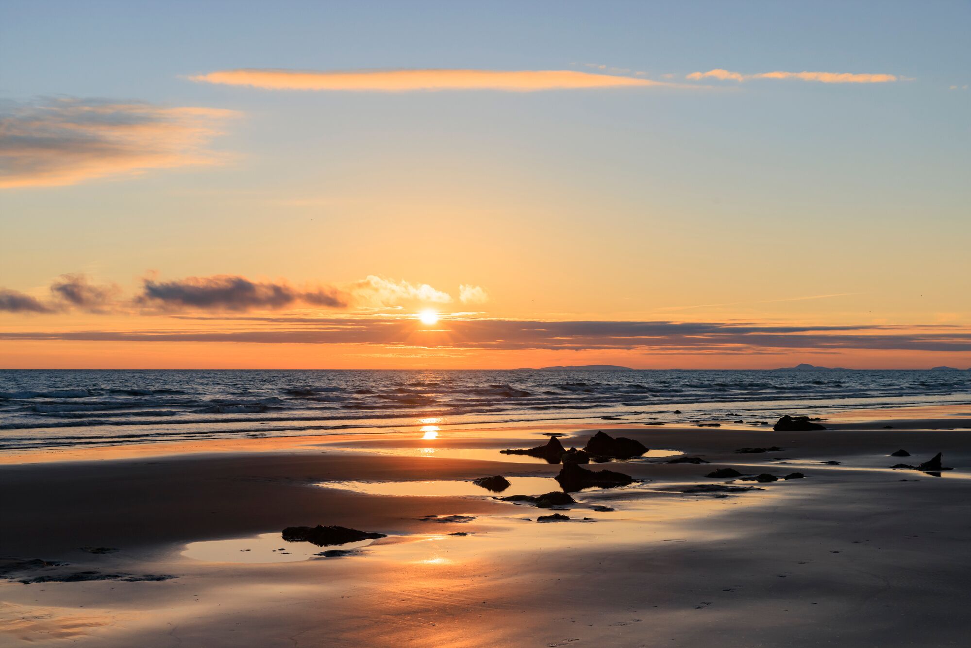 A beach at sunset with calm waves and scattered rocks.