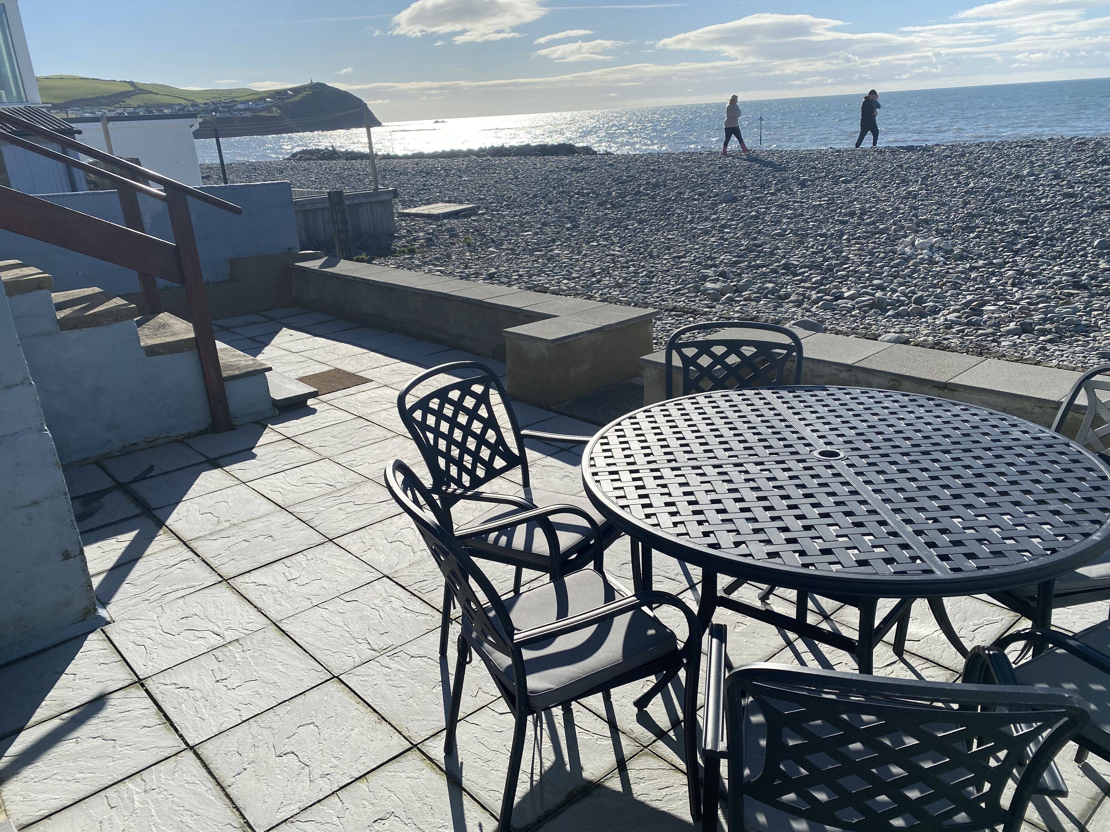 A terrace with a table and chairs, overlooking a pebble beach and the sea.