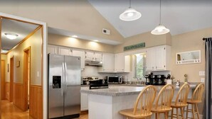 A modern kitchen with white cabinets, a stainless steel refrigerator, a microwave, and a coffee maker. There is a bar area with wooden stools. - Oceanside 7BR House with Private Pool and Hot Tub (Corolla)