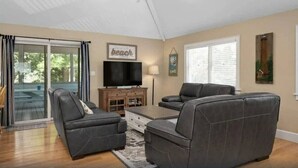 A living room with a television, two grey sofas, a coffee table, and a sliding glass door leading to an outdoor area. - Oceanside 7BR House with Private Pool and Hot Tub (Corolla)