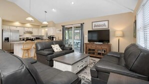 A modern living room with a sofa, coffee table, and a television. There is a kitchen area with cabinets and appliances visible. - Oceanside 7BR House with Private Pool and Hot Tub (Corolla)