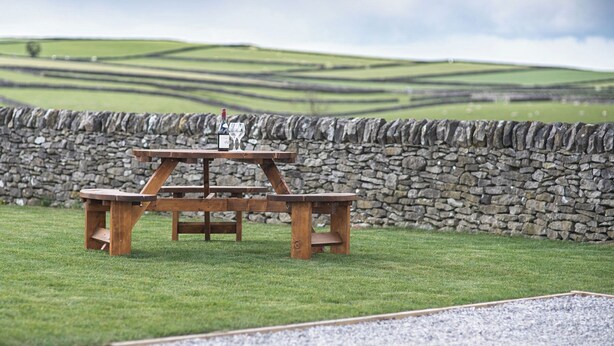 A picnic table with a bottle and glass on it, set against a stone wall and grassy field with rolling hills in the background.