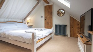 A bedroom with a sloped ceiling, a bed with white linens, a bedside lamp, a wooden door, a round mirror, a radiator, and a wooden dresser with a remote control.