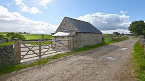 A stone farmhouse with a metal gate, a dirt road, and green fields in the background.
