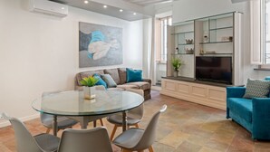 A modern living room with a glass dining table, grey chairs, a sofa with blue cushions, a mounted TV, and a fireplace. - ViaPaola43 Palace 1st floor 2Bdr (Roma)