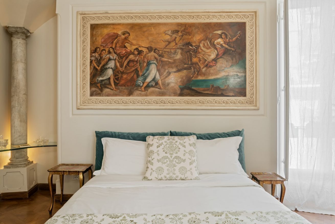 Une chambre à coucher avec un grand tableau de style classique, un lit avec une literie blanche et verte, et une table en verre sur laquelle est posé un vase.