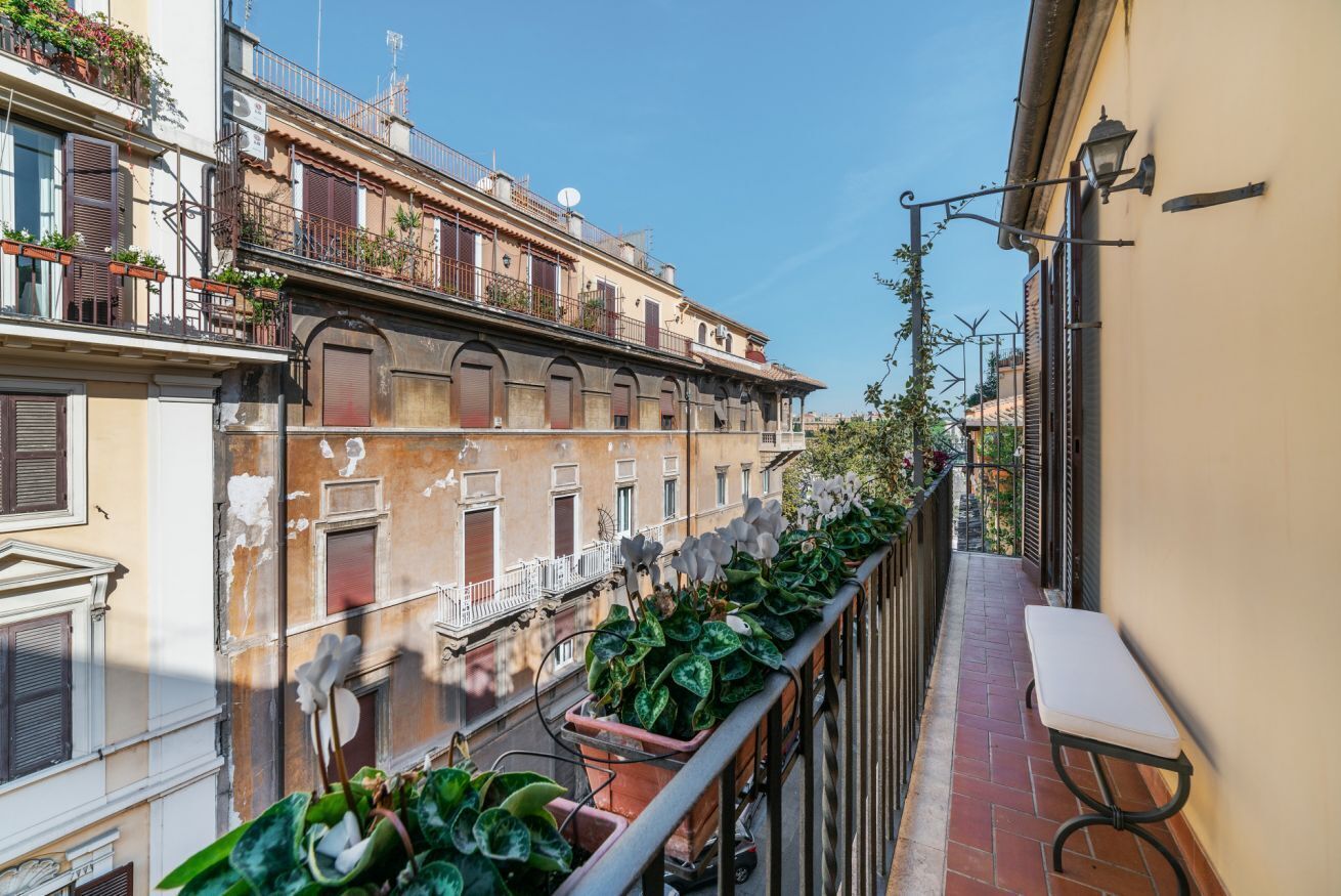 A narrow urban street with buildings on either side, a balcony with potted plants, and a bench.