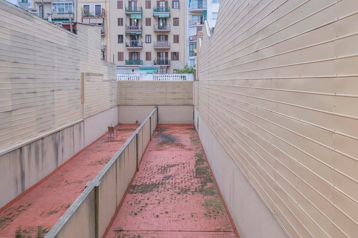 Un couloir étroit au sol en briques rouges, bordé de murs de couleur beige, qui mène à un balcon doté d’une balustrade blanche.
