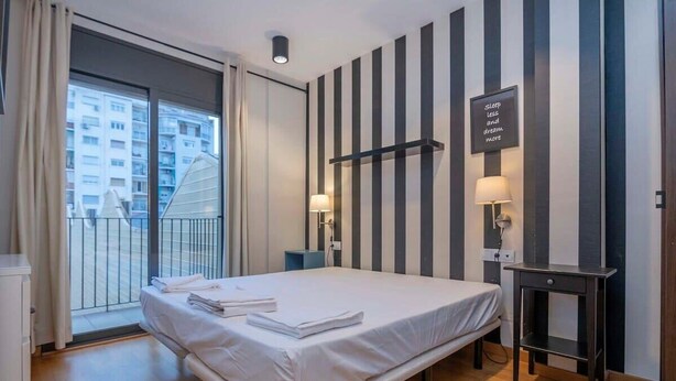 A hotel room with a bed, two bedside tables with lamps, a black and white striped wall, and a view of buildings through a large window. - Eixample 2 bedroom with private terrace (Barcelona)