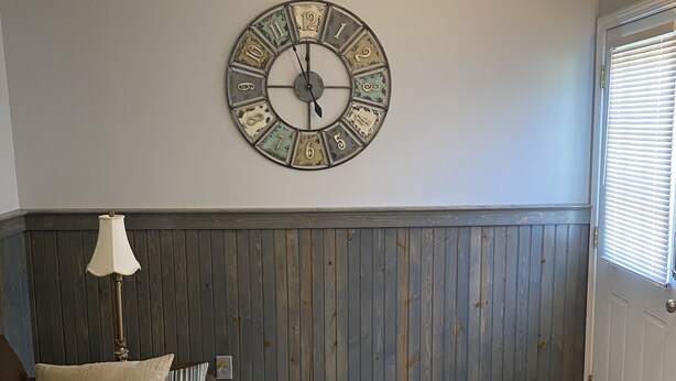Een kamer met een ronde wandklok, een houten lambrisering, een bruine bank met kussens, een vloerlamp en een deur met zonwering.