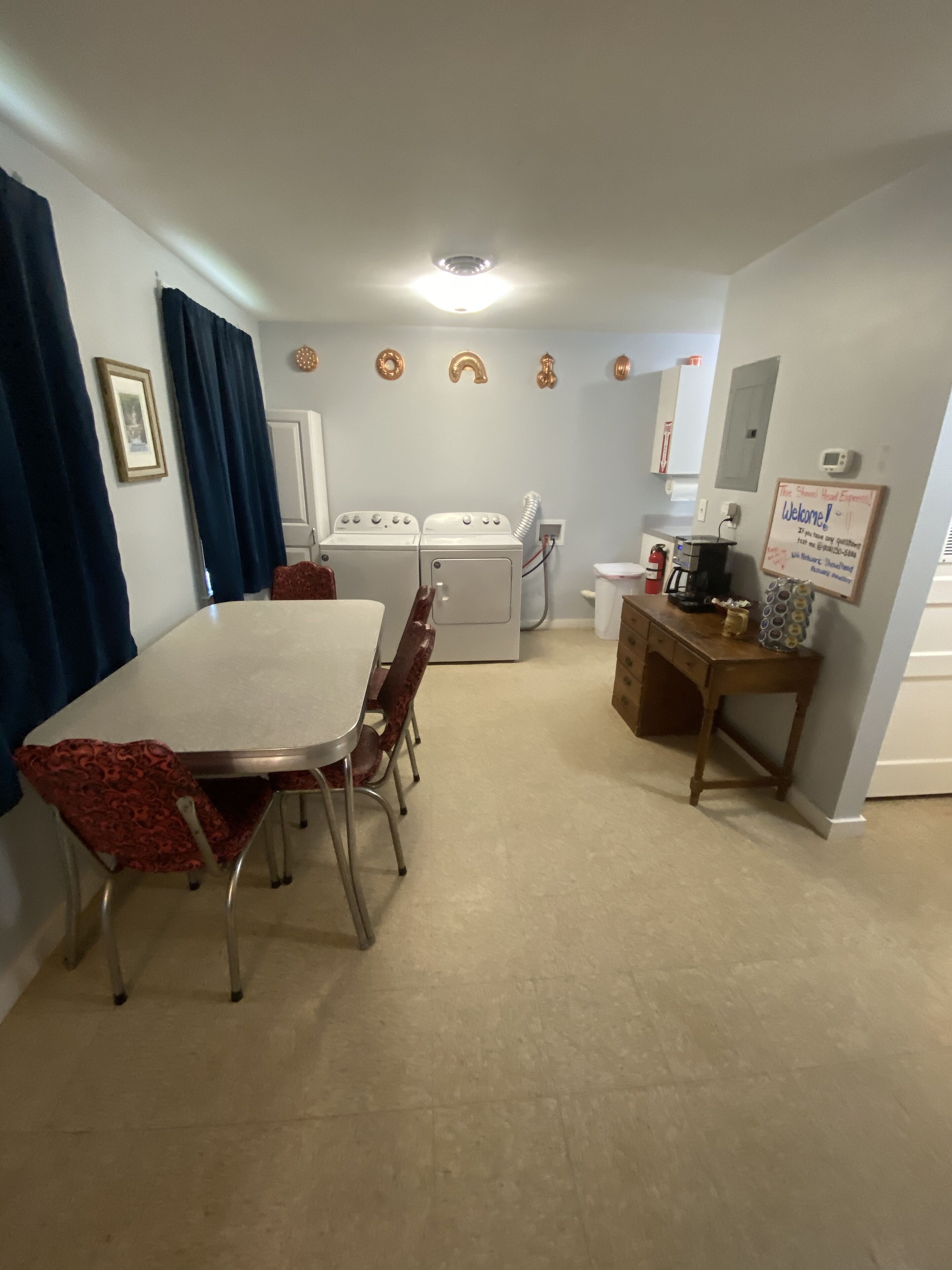 Une pièce comprenant une table à manger et des chaises, un lave-linge, une machine à café et un panneau accroché au mur.