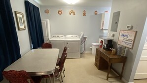 Een kamer met een eettafel en stoelen, een wasmachine, een koffieapparaat en een bordje aan de muur.