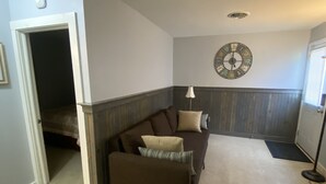 Een woonkamer met een bruine bank, een klein voetenbankje, een wandklok en een deur die naar een andere kamer leidt.