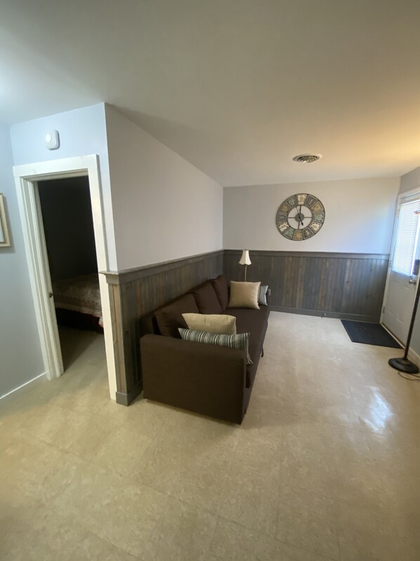 A living room with a brown sofa, a small ottoman, a wall clock, and a door leading to another room.