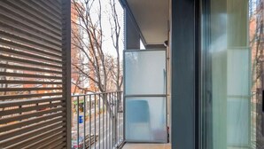 Un balcón con barandilla de metal, puerta de vidrio y vista a una calle con coches y árboles.