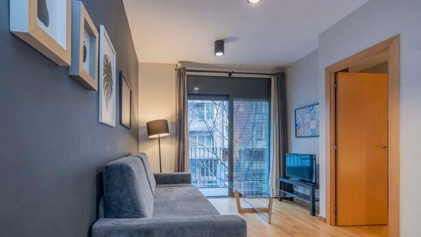 Una sala de estar moderna con un sofá gris, un televisor sobre un mueble bajo y un ventanal con persianas.