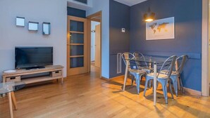 A modern living room with a flat-screen TV on a wooden stand, a glass table, and a dining area with metal chairs. - Plaza España 2 bedroom with balcony (Barcelona)