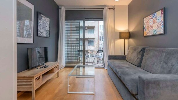 Ein modernes Wohnzimmer mit grauem Sofa, einem Glastisch, einem Fernseher auf einem Holzständer und Kunstwerken an den Wänden.