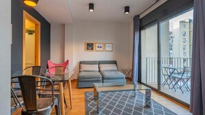 Ein modernes Wohnzimmer mit einer Couch, einem Glastisch und einem Essbereich mit Stühlen und Tisch. An den Wänden hängen Bilder und der Balkon ist mit Gartenmöbeln ausgestattet.