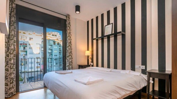 Ein Hotelzimmer mit einem großen Bett, einem kleinen Nachttisch, einer an der Wand befestigten Ablage und Blick auf Gebäude durch eine Glastür.