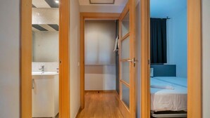 Ein schmaler Flur führt zu einem Badezimmer mit Waschbecken und Dusche. Im angrenzenden Zimmer befindet sich ein Bett mit blauer Kopfteil.