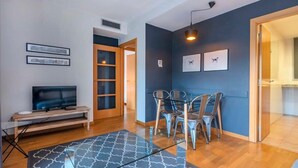 Ein modernes Wohnzimmer mit einem Flachbildfernseher auf einem Holzständer, einem Glastisch mit Metallstühlen und einem gemusterten Teppich.