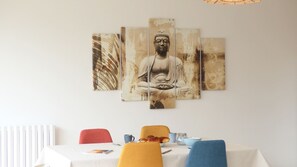 A dining area with a Buddha painting, a table with a white tablecloth, and chairs in red, yellow, and blue.