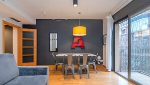 Un moderno salón con un sofá gris, una mesa de comedor con sillas, un ventanal con vistas a edificios y una lámpara colgante amarilla.