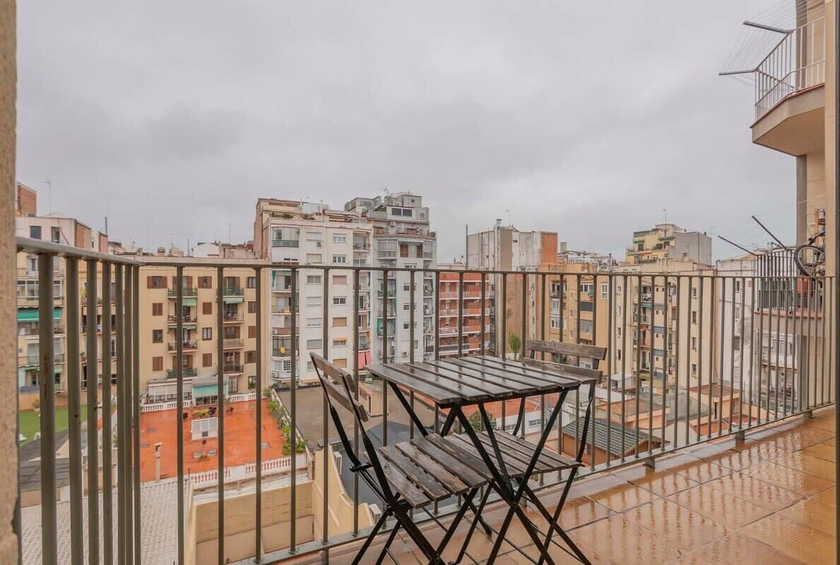 Ein Balkon mit Metallgeländer, einem Holztisch und zwei Stühlen. Im Hintergrund sind mehrere Wohngebäude zu sehen.