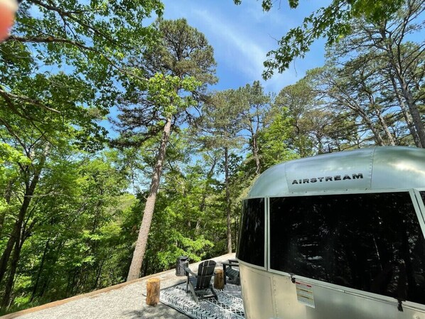 Una caravana con la marca 'AIRSTREAM' estacionada en una zona boscosa con árboles y cielo despejado.