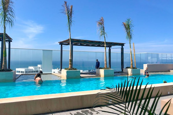 A rooftop infinity pool with palm trees, a pergola, and a clear view of the ocean.