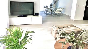 A modern living room with a flat-screen TV, a dining area with a glass table and metal chairs, and a large potted plant. - 2 Coastal villas 5 min. From beach. Panoramic view of sea and sunset (Καλύβια Θορικου)
