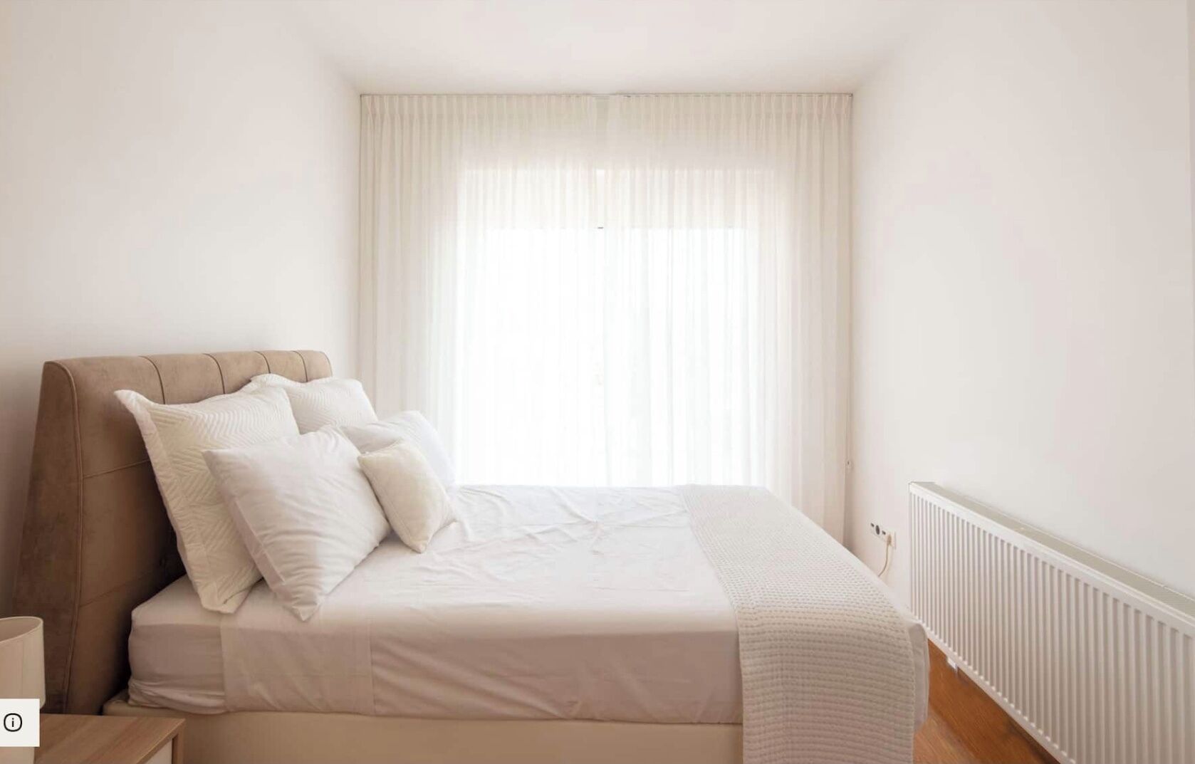 A bedroom with a bed, pillows, a radiator, and sheer curtains.