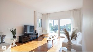 A modern living room with a flat-screen TV, a sofa, a coffee table, and a view of greenery through large windows. - 2 Coastal villas 5 min. From beach. Panoramic view of sea and sunset (Καλύβια Θορικου)