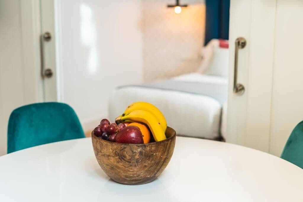 Un cuenco de madera con frutas sobre una mesa blanca en una habitación con sillas color turquesa y un sofá blanco.