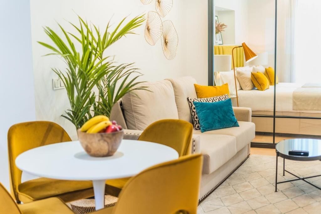 A modern living room with a sofa, a coffee table, and a dining area with chairs and a table. There is a plant and a bowl of fruit on the dining table.