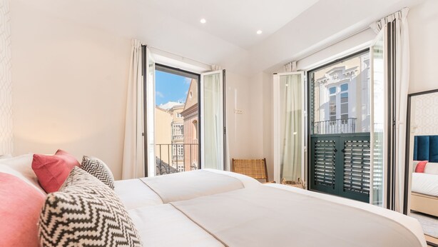 A hotel room with a large bed, a chair, and a view of the city through the windows. - Debambú Suites (Málaga)