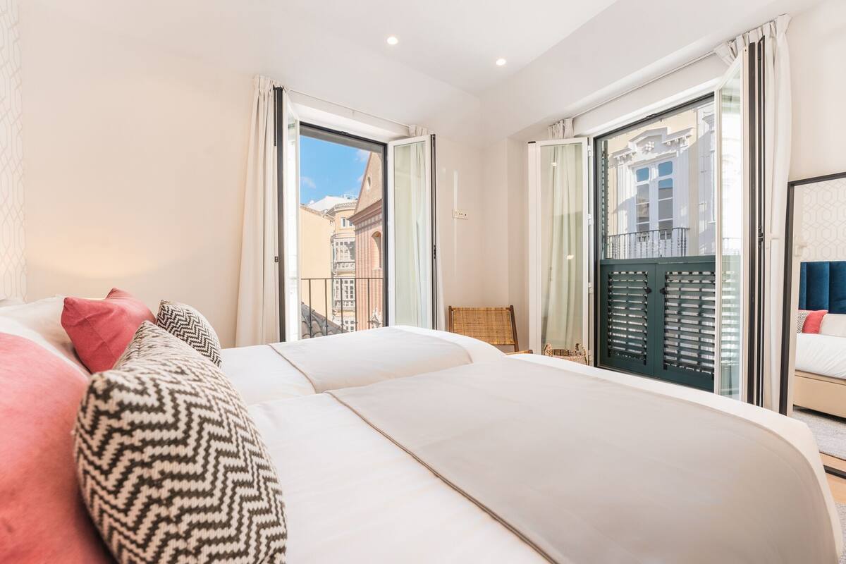 Quarto de hotel com uma cama grande, uma cadeira e vista para a cidade pelas janelas.