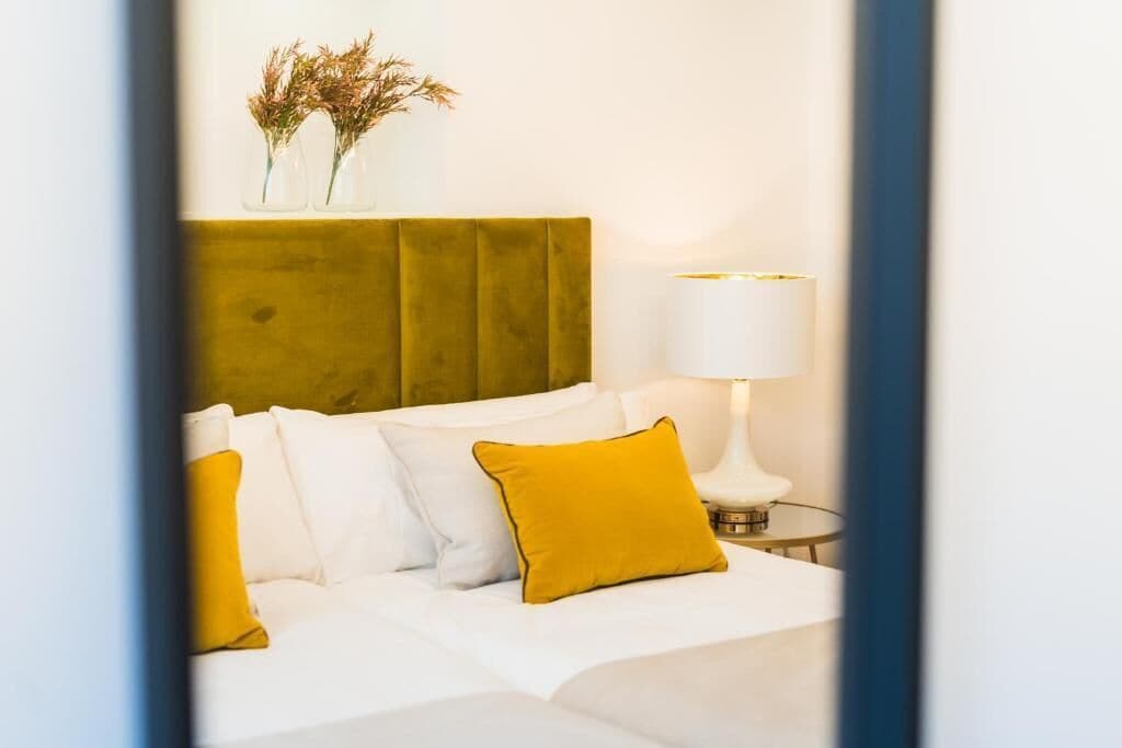 A neatly made bed with white and yellow pillows, a green upholstered headboard, and a bedside table with a lamp.