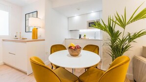A modern kitchen with white cabinets, a microwave, and a sink. There is a dining table with yellow chairs and a bowl of fruit. - Debambú Suites (Málaga)
