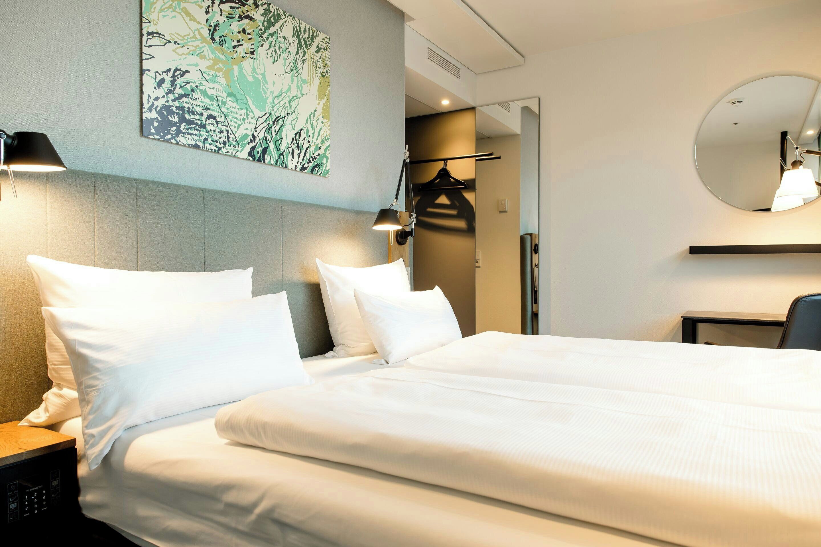 Ein Hotelzimmer mit Bett, Nachttischlampen, einem Spiegel, einem Kleiderständer und einem Wandbild.