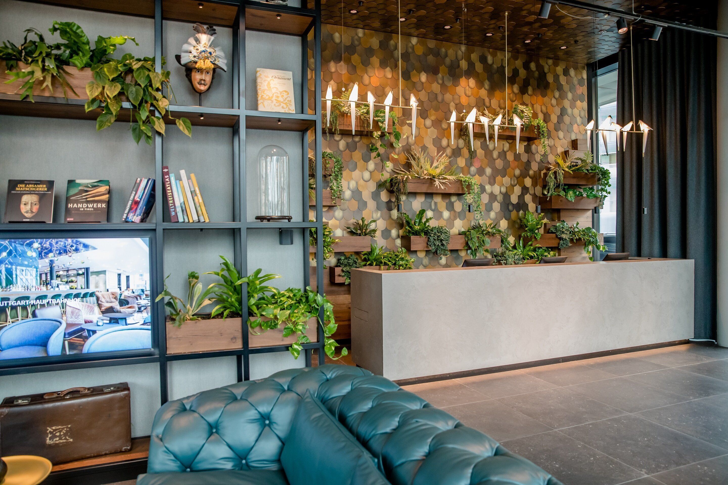 Uma área de recepção moderna com um sofá, prateleiras com livros e objetos decorativos, uma grande janela com vista para a cidade e uma parede decorada com plantas e velas.
