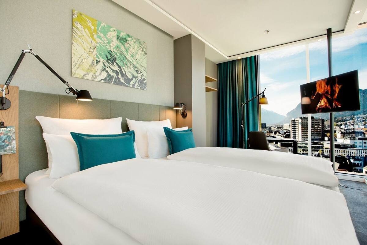 A hotel room with a large bed, a city view, a painting on the wall, and a TV displaying a fire.