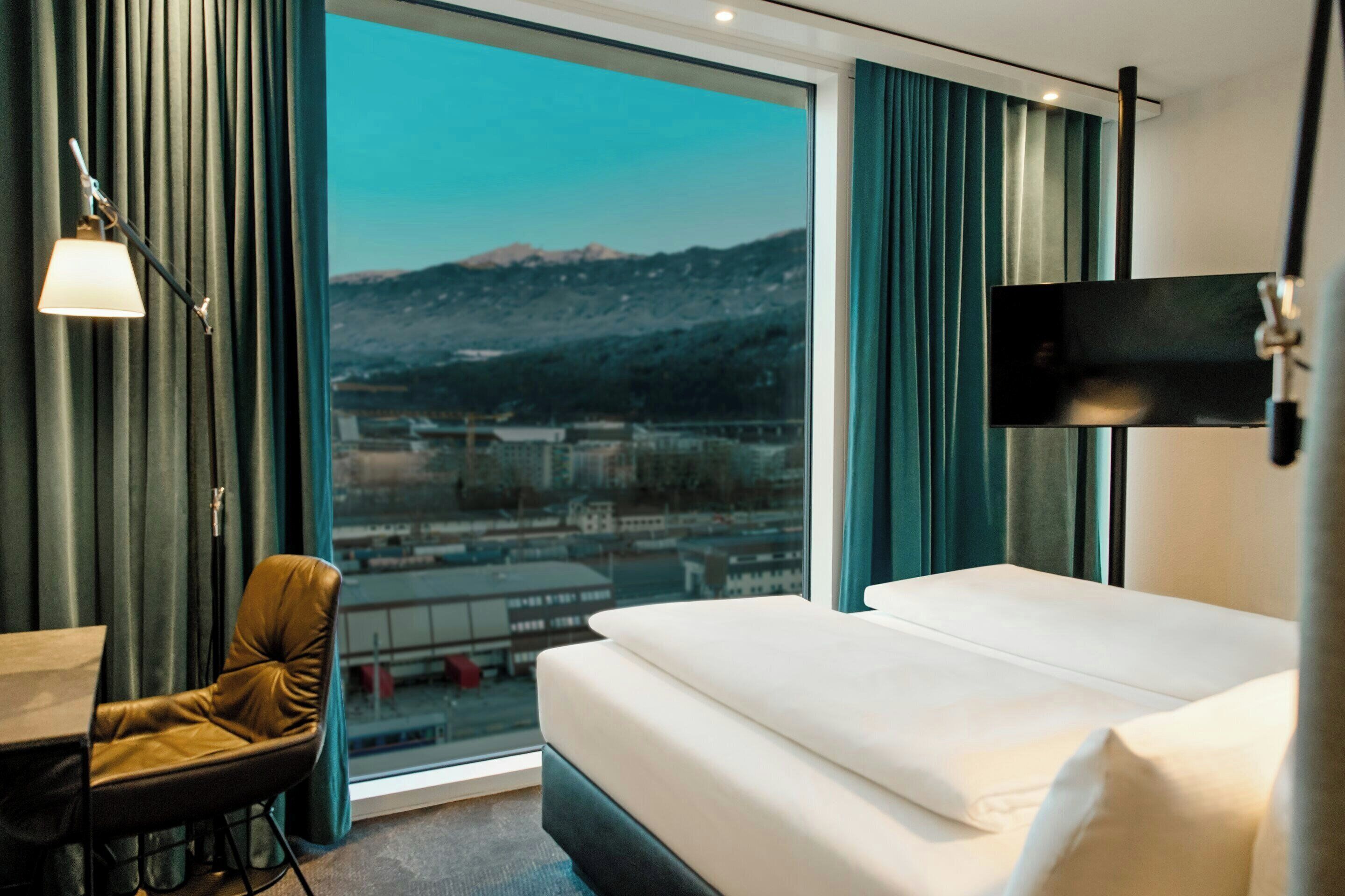 A hotel room with a bed, a desk, a chair, a TV, and a large window with a view of a city and mountains.